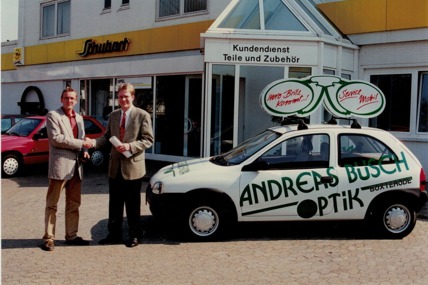 Zwei Männer stehen vor einem Kundendienstgebäude neben einem mit Andreas Busch Optik beschrifteten Kleinwagen.