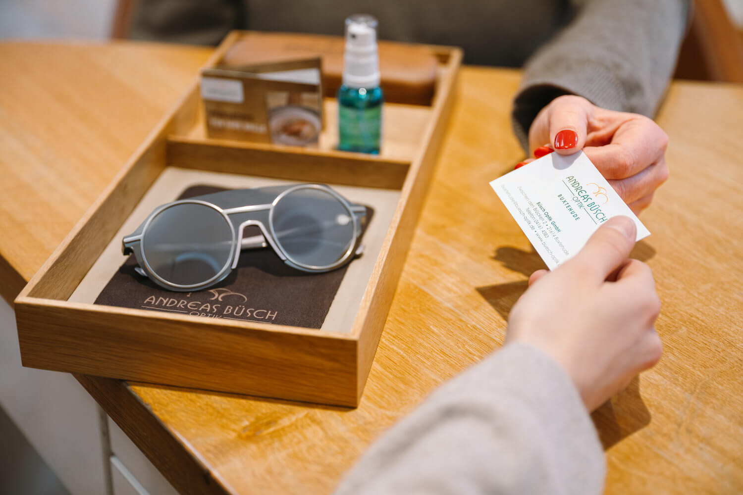 Hände überreichen eine Karte; daneben liegen eine Sonnenbrille, ein Spray und weitere Produkte in einer Holzbox.