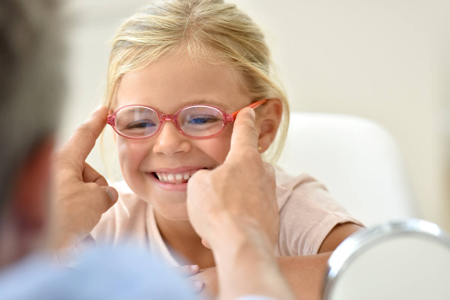 Lächelndes Mädchen mit roter Brille, deren Sitz ein Erwachsener anpasst.