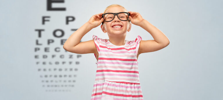 Mädchen mit Brille hält sich vor einer Sehtafel die Hände an den Brillenbügeln.