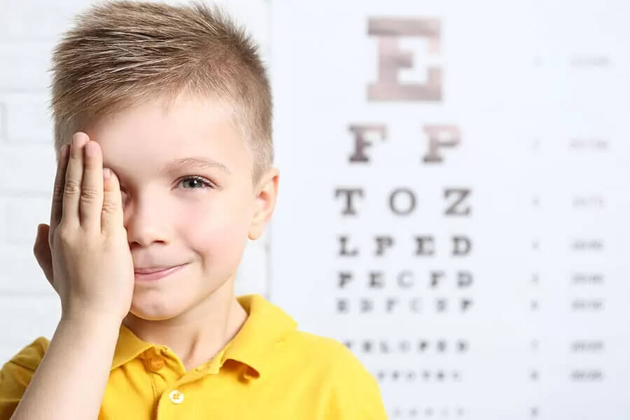 Junge hält sich bei einem Sehtest ein Auge zu und blickt in die Kamera; dahinter ist eine Sehtesttafel zu sehen.