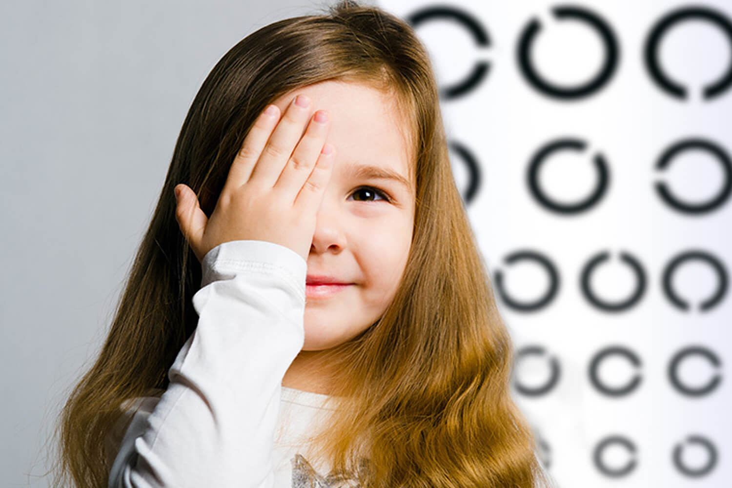 Mädchen hält sich beim Sehtest ein Auge zu, im Hintergrund ist eine Sehtesttafel mit unterschiedlich großen Ringen zu sehen.