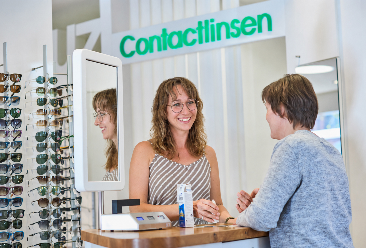 Zwei Frauen in unserem Optikgeschäft im Gespräch an einem Verkaufstresen, daneben ein Spiegel und ein Regal mit Sonnenbrillen.