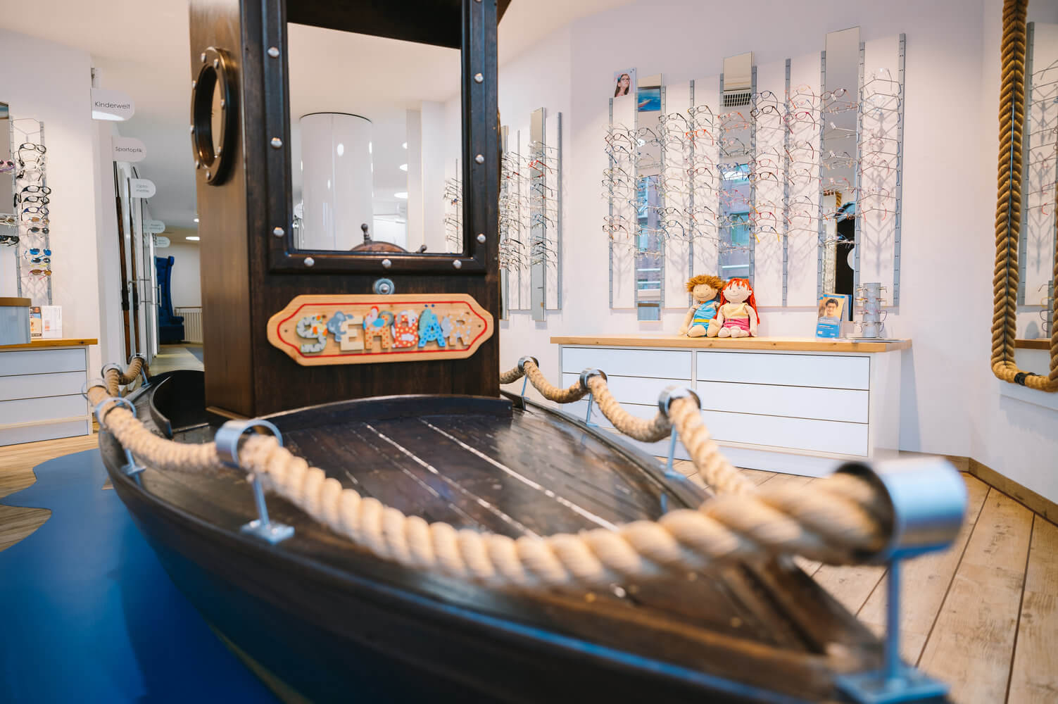 Historisches Boot mit Seilumrandung und schwarzem Aufbau in einer hellen Ausstellung mit Brillenregalen und Puppen.
