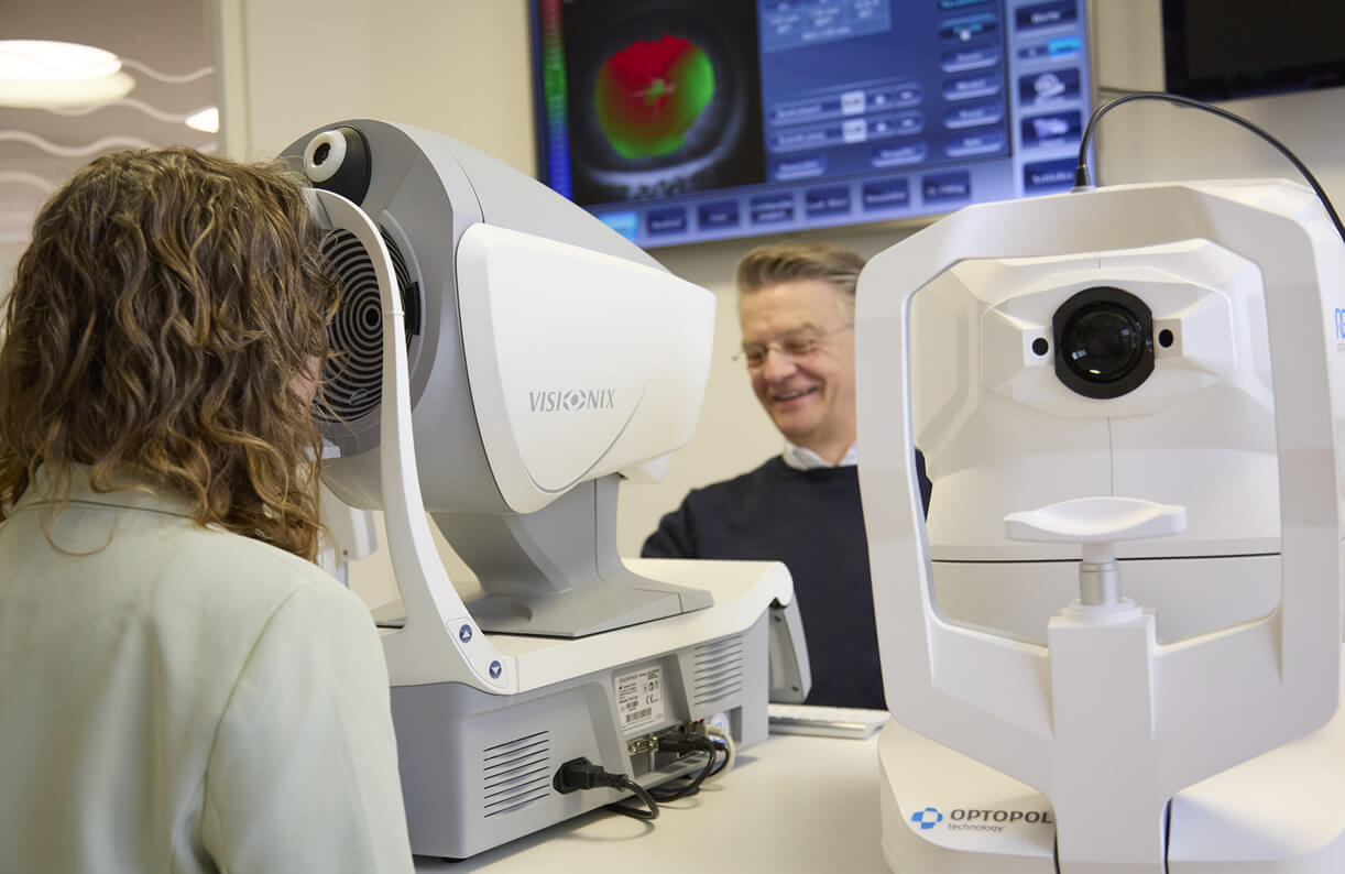 Augenuntersuchung am Messgerät und einem Optometristen im Hintergrund.