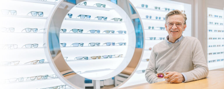 Optiker steht lächelnd vor einer Wand mit Brillengestellen und hält eine Brille in der Hand.