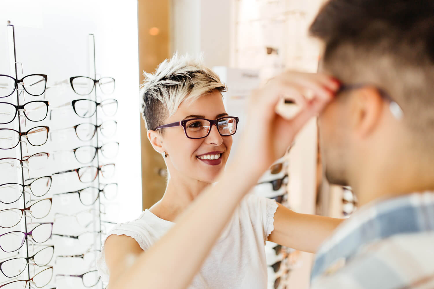 Eine Person probiert in einem Optikergeschäft eine Brille an, während eine andere Person sie im Spiegel betrachtet.