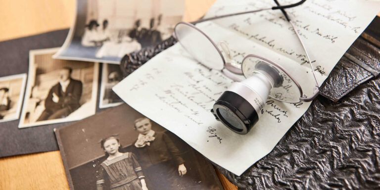 Historische Fotografien, handschriftliche Dokumente, eine Lupe und ein altes Lederetui auf einem Tisch.