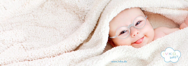 Baby mit rosafarbener Brille liegt eingekuschelt in einer weißen Decke; rechts unten sind ein Logo und eine Webadresse sichtbar.